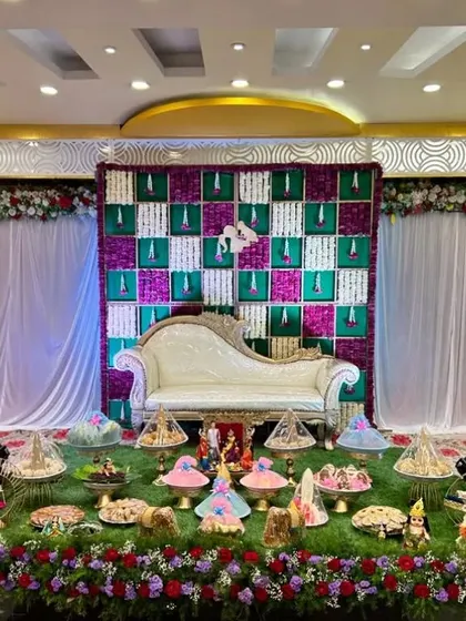 A Seemantham ceremony with a checkered backdrop of purple and green flowers and an elaborate plate display on an artificial grass floor.