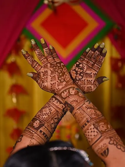 A bride showing off her full bridal mehendi, with both hands raised against a colorful mehendi function background.