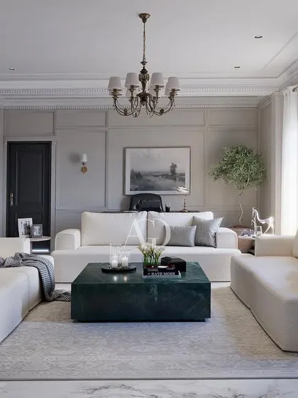 An example of "soft minimalism" where classical principles are applied with a restrained hand. The room features clean, modern sofas and a striking green marble coffee table, all framed by traditional wall paneling and a classic chandelier.