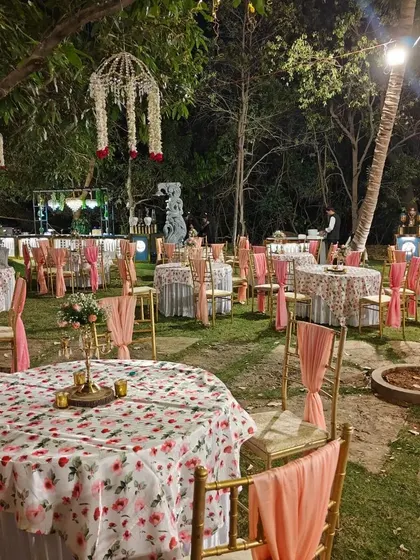 An enchanting outdoor reception setup amidst the lush greenery of the ashram. The floral tablecloths, elegant chair drapes, and hanging chandeliers create a magical atmosphere for an evening celebration.