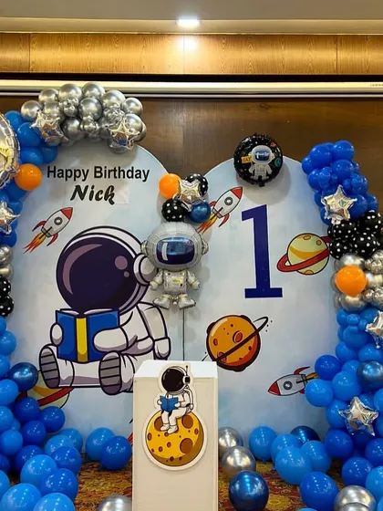The main backdrop for a space-themed first birthday. The decoration includes a large astronaut cutout, a moon foil balloon, and a blue and silver balloon arch representing the galaxy.