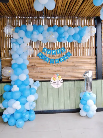 A simple blue and white L-shaped balloon arch for a first birthday against a bamboo wall.