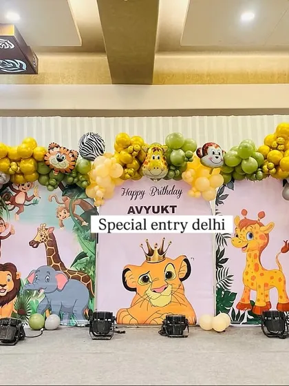 A Lion King inspired jungle theme backdrop for a first birthday. The setup features characters like Simba, Timon, and Pumbaa, along with a balloon arch and PAR lights to illuminate the stage.