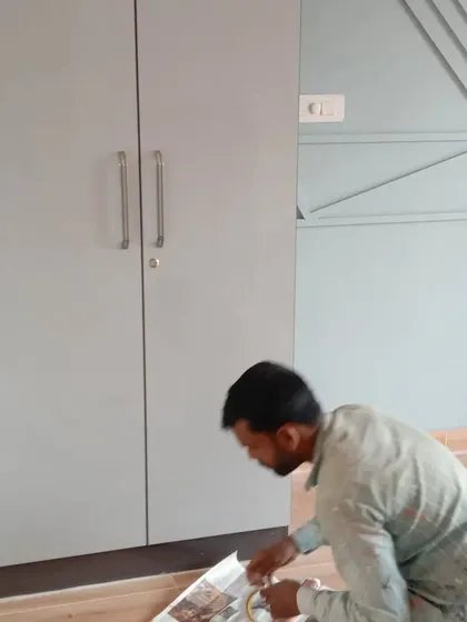 A painter applies masking tape along the floor edge next to a wardrobe. This ensures a sharp, clean line between the wall and the floor, and protects the flooring from paint.