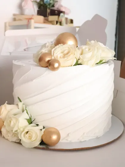 A gorgeous all-white cake with a diagonal textured frosting, decorated with white roses and gold spheres.