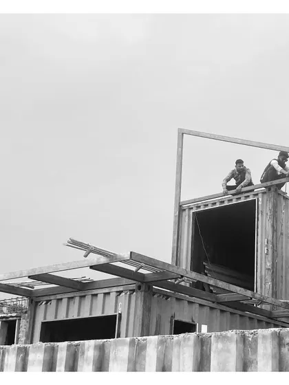 Workers begin fabricating the framework on top of the stacked containers. This black and white photo captures the hands-on process of building a custom structure.