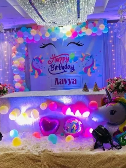 A magical unicorn-themed birthday stage. The backdrop features unicorn illustrations and is framed by pastel rainbow balloons, with a light-up unicorn prop and fairy lights.
