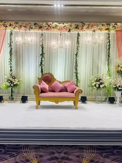 A simple yet elegant stage design for a Roka or engagement. A pink sofa is set against a backdrop of white and pink drapes, with hanging chandeliers and floral vines adding a touch of romance.