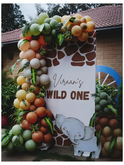 A 'Wild One' welcome sign framed by a unique balloon garland with green and orange tones, accented with faux leaves. A cute elephant cutout completes this jungle party entrance.