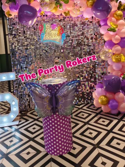 A full view of the purple butterfly theme, showing the arrangement of the balloon arch, the light-up number '5', and the butterfly-wing cake stand in front of a silver sequin wall.
