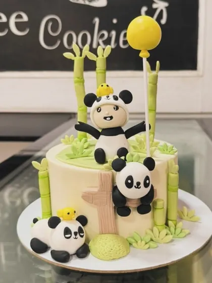 An adorable panda-themed cake for a first birthday. The design includes multiple cute panda figures, bamboo stalks, and a child in a panda costume.