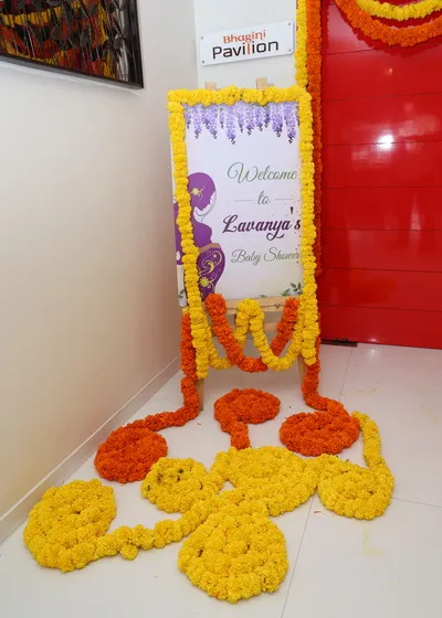 A welcome sign for a baby shower, framed with yellow and orange marigold garlands. The floral arrangement on the floor adds a traditional and welcoming touch.