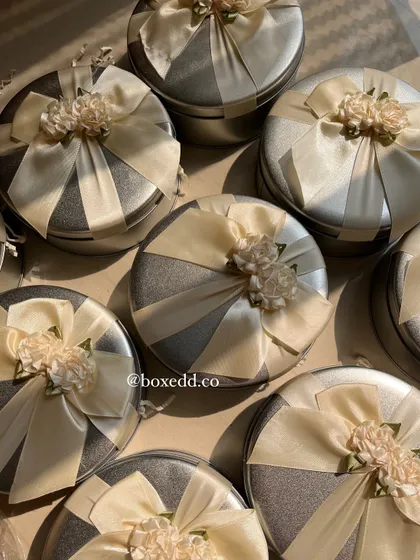 Beautifully decorated tin boxes make for wonderful return gifts. These silver tins are adorned with satin ribbons and floral embellishments, ready to be filled with sweets, candles, or other small favors.