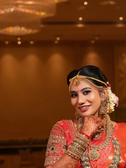 A happy bride posing for a portrait in a grand convention hall, her red saree vibrant against the warm lighting.