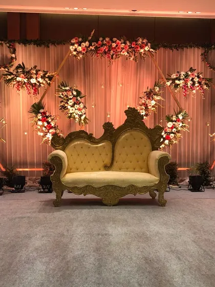 A classic stage decor for a reception or engagement. A vintage-style golden sofa is set against a backdrop of drapes and a geometric frame adorned with flowers.