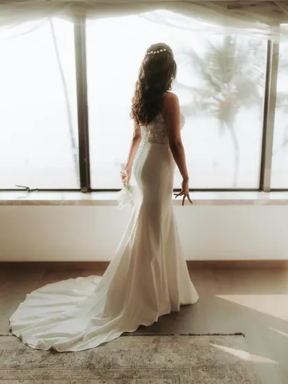 A beautiful portrait of a bride in her white gown, looking out a large window with palm trees in the background.