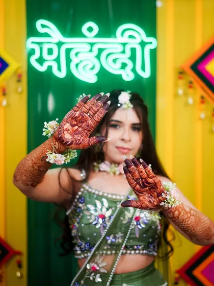 A beautiful bride-to-be posing in front of a neon "Mehndi" sign. Her hands showcase a rich, dark henna stain, complemented by floral jewelry for the perfect sangeet look.