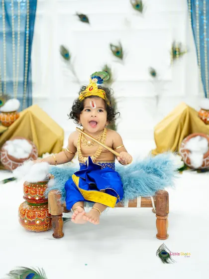 A mischievous little Krishna sitting on a cot with his flute. The falling peacock feathers add a dynamic and magical touch to this Janmashtami photo.