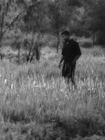 A black and white environmental portrait of a man standing in a field of tall, sunlit grass. The soft focus and natural setting give the image a calm and thoughtful quality.
