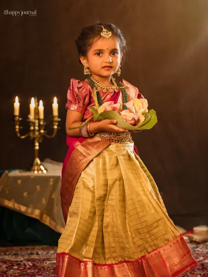 A little girl in a traditional silk lehenga stands holding a lotus flower offering, with the soft glow of candles in the background.
