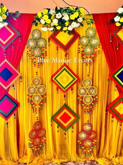 A vibrant Haldi and Mehendi backdrop with red and yellow drapes and colorful traditional hangings.