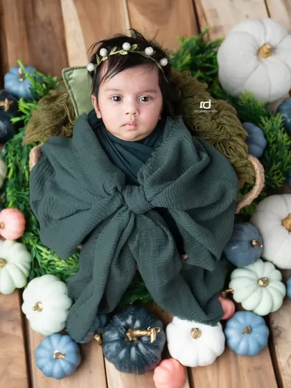Our little pumpkin, wide-eyed and curious, wrapped in a green bow and surrounded by colorful pumpkins.