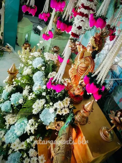 A close-up of the Krishna statue at a devotional event. The statue is surrounded by beautiful flowers, and the background features a peacock feather motif, which is strongly associated with Lord Krishna.