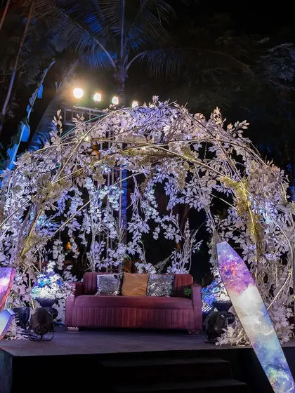 A cozy seating area at the celestial sangeet. The sofa is placed under an arch of silver leaves, with glowing crystal props on either side, creating a magical, enchanted forest feel.