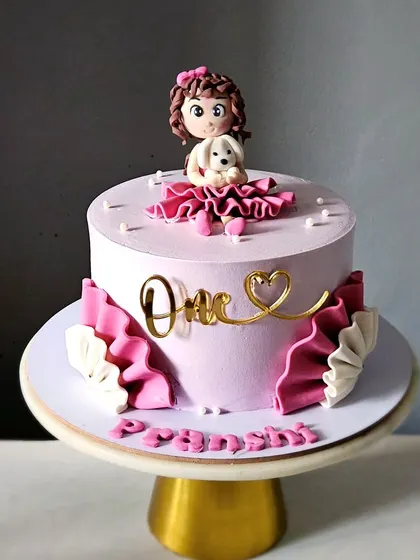 A pretty pink cake for a first birthday, featuring a handcrafted girl figurine holding her puppy. The cake is decorated with elegant fondant ruffles.