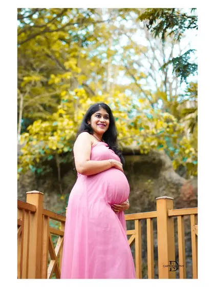 A full-length portrait of an expecting mother in a pink gown, framed by the natural beauty of a park with yellow-leaved trees.