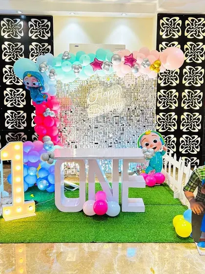 A Cocomelon first birthday party with a silver shimmer wall and a "ONE" table on a green turf floor.