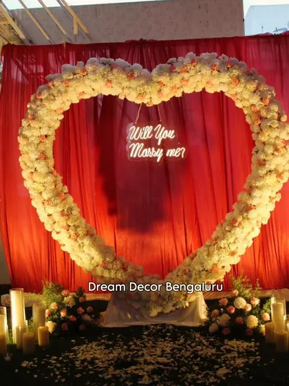 A romantic proposal setup with a large heart-shaped arch made of white and pink flowers. The 'Will You Marry Me?' neon sign and candle-lit path create an unforgettable moment.