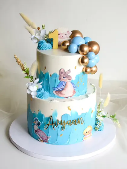This Peter Rabbit themed cake was for a first birthday celebration. The two-tiered design has a blue watercolor effect and is decorated with edible character prints, gold and blue spheres, and dried grasses.