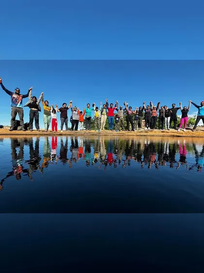 A large group of trekkers celebrating by a stunning mountain lake, their reflection perfectly mirrored in the water.
