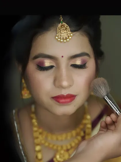 A close-up of the bridal makeup process for Sheela's muhurtham. This shot highlights the application of a bold red lip and shimmery gold eyeshadow, key elements of a classic bridal look.