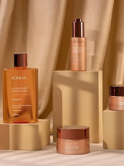 A full range shot of Koheva's skincare products. The warm, earthy tones, geometric blocks, and draped satin cloth create a luxurious and elegant composition, with a window-like shadow adding depth.