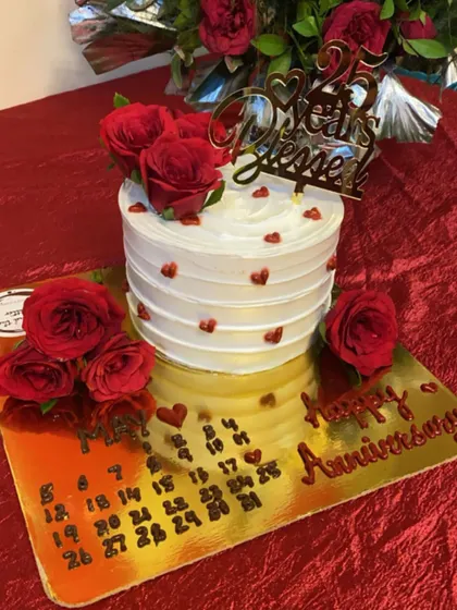 A heartfelt cake for a 25th anniversary. This design features a "25 Years Blessed" topper, fresh red roses, and a custom calendar marking the special date on the cake board.