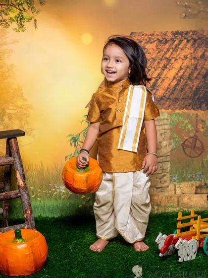 A happy little boy in a traditional dhoti and kurta, holding a pumpkin in a rustic village setting. His joyful expression perfectly captures the fun of our themed sessions.