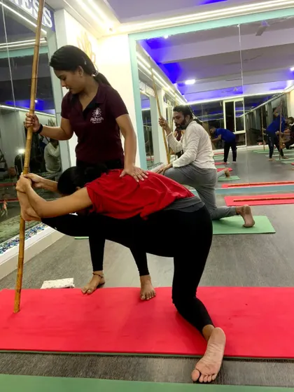 Using a yoga stick for leverage, an instructor helps a student deepen their stretch in a variation of Hanumanasana (split pose).