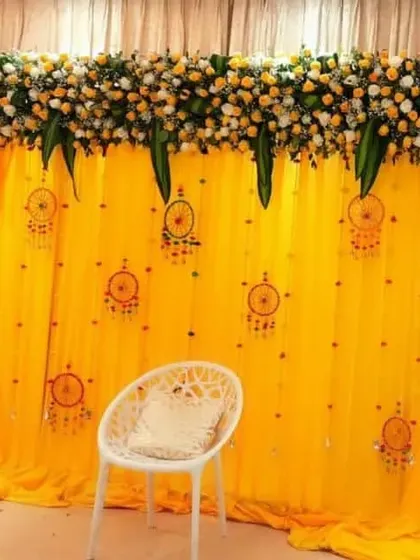 A bright and cheerful Mehandi backdrop with a yellow curtain, decorated with dreamcatcher hangings and a floral border at the top.