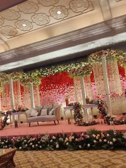 A large and lavish stage setup for a grand reception. The backdrop is a sea of red and pink flowers, with hanging floral strings and extensive arrangements lining the entire length of the stage.