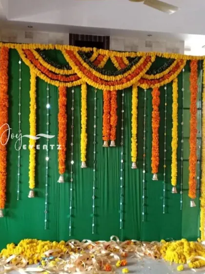 This backdrop for a half saree ceremony features a dark green fabric with hanging garlands of yellow and orange marigolds. We added strings of pearls and small bells for a delicate and festive look.