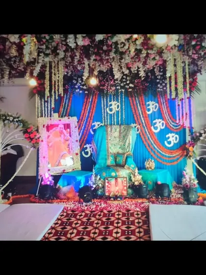 This Guruji Satsang stage decoration was designed with a vibrant blue color theme as requested by the client. The setup includes a floral canopy, a draped backdrop with marigold 'Om' symbols, and a decorated Aasan.