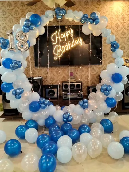 A stylish 18th birthday decoration for home, with a blue and white balloon ring, neon sign, and fairy lights set up around a TV unit.