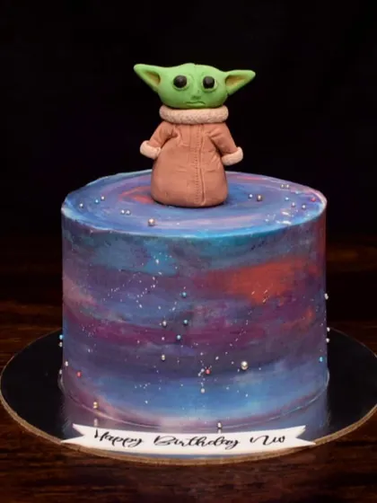 This is the way. A galaxy-themed cake featuring a hand-molded fondant Baby Yoda (Grogu) on top. The buttercream base is painted with cosmic colors to create a Star Wars cake that's out of this world.