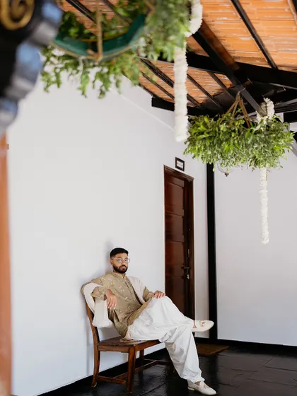 A stylish portrait of the groom, seated in a quiet corner of the venue under hanging greenery.