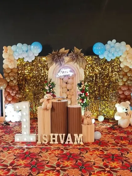 A cozy bear-themed birthday setup with a gold sequin backdrop. The decor uses a warm color palette of brown and blue, with cute teddy bears and a light-up number '1'.