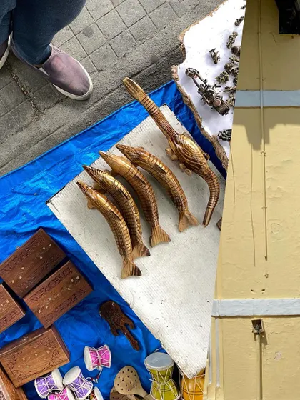A collage of street finds. Carved wooden fish and tiny drums from a market stall are paired with the simple, yellow facade of a building, creating a playful composition.