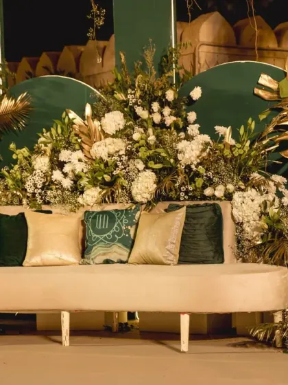 A close-up of the reception stage seating, showing the plush cream sofa against a rich green backdrop, surrounded by lush white florals and gold accents.