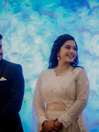 The couple stands on stage at their Sangeet, dressed in elegant formal wear, ready to celebrate with their loved ones.